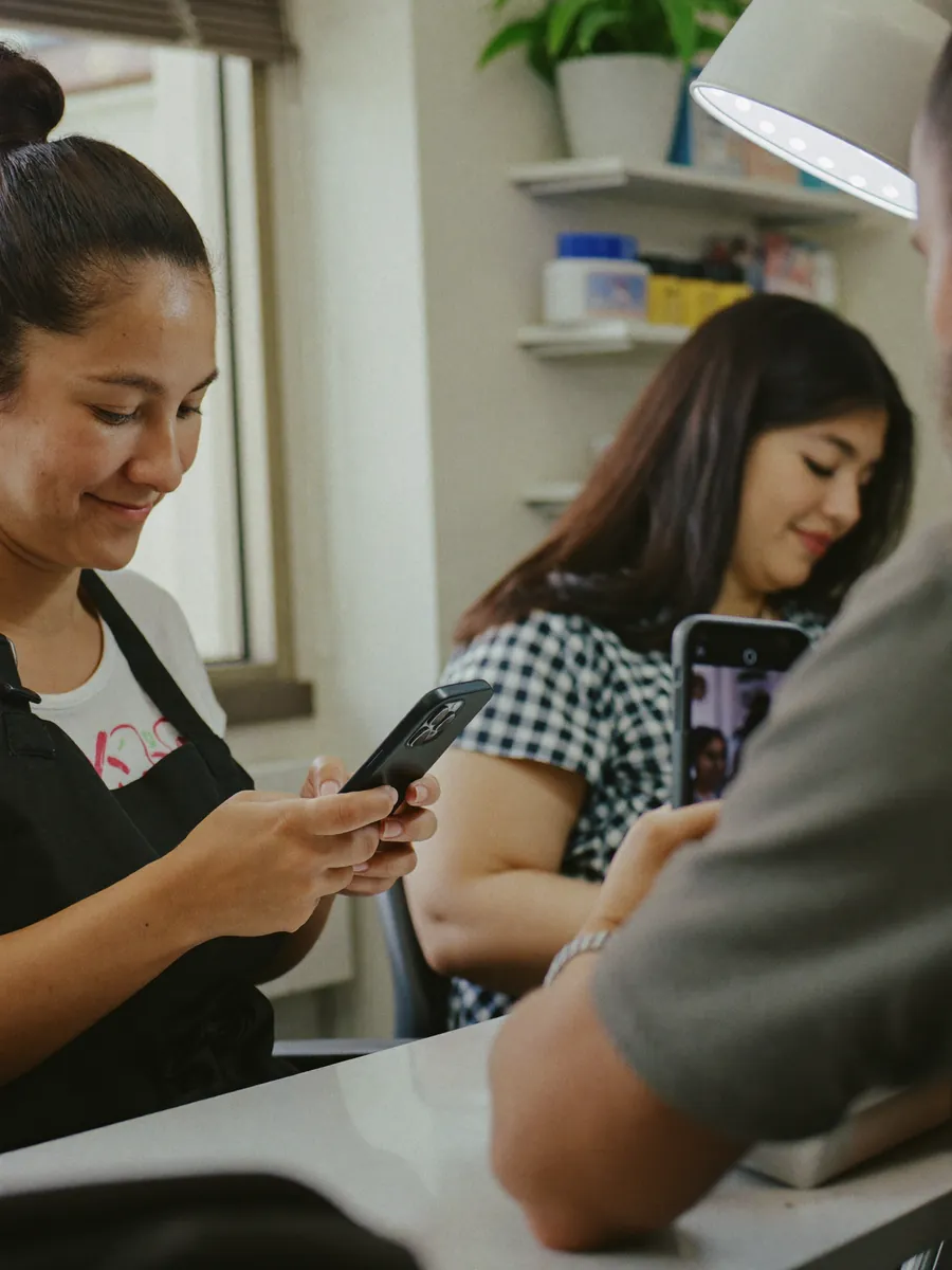 Mulher de avental no salão onde trabalha sorri olhando o celular entre um atendimento e outro.